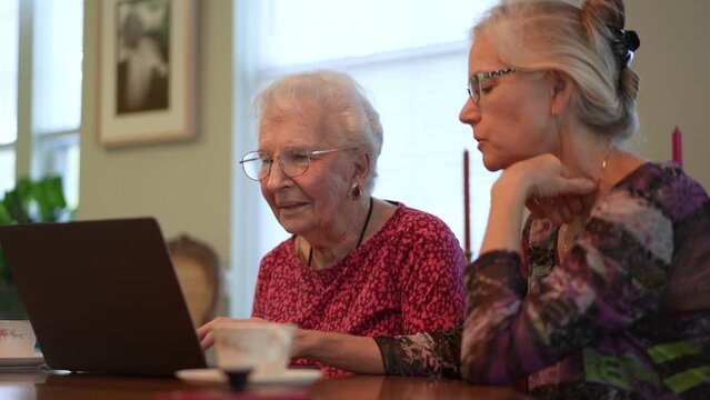 Happy elderly middle aged woman sitting at table with grown up daughter, watching family or journey photos on computer. Smiling blonde lady listening to older, using laptop.