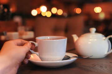 Closeup of female hand with natural manicure holding cozy ceramic white mug of tea or coffee. Relax and comfort at home, cafe. Empty space for text on blurry background, backdrop
