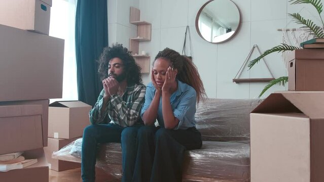 Young multiracial sad African American woman and Arabian man frustrated by having to move out of house after payment delays at mortgage or rent sit on sofa surrounded by cardboard boxes