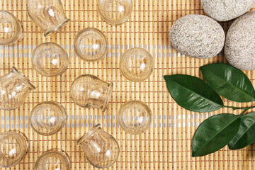 Glass cups for cupping therapy on a bamboo mat with stones and a green twig, flat lay composition.