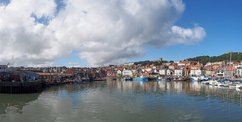 Fototapeta na wymiar long panoramic view of the harbour in scarborough on a summer day with fishing boats reflected in the water and town buildings