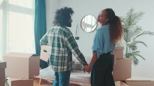 Young love couple African American woman and Arabian man enters new apartment holding hands and hugs excitedly after relocation standing among cardboard boxes. Multiracial lifestyle