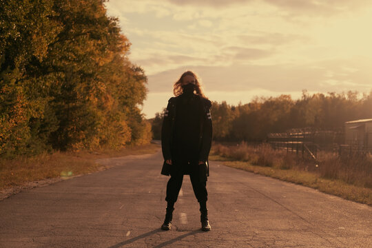 Young stylish man with long hair at sunset standing on the road wearing a black mask during pandemic