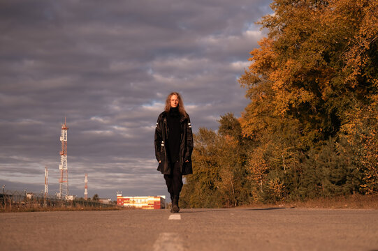 Young male blond model with long hair wearing black raincoat walking in the rays of sunset along the road outside city