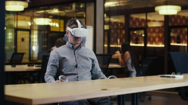 Male office worker wearing VR headset and using wireless controllers, gesturing, watching data in virtual reality. Work in office of hi-tech company near coworkers. Cyberspace digital technology