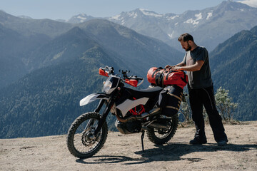 motorcyclist fastens dry bag on the trunk of his off-road motorcycle in incredibly beautiful mountainous summer landscape