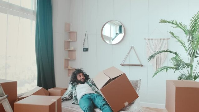 Young clumsy positive Arabian man millennial holds lot of cardboard boxes for move and falls on bed with smile rejoice relocation posing in new house or apartment. Mortgage, moving concept