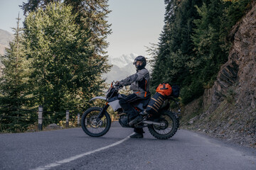 Biker man standing on dirt motorcycle loaded with big dry bag and wardrobe trunks on asphalt mountain road