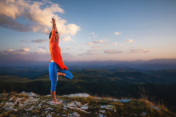 young woman in a tree pose outdoors in the mountains watching the sunset, yoga balance near the cliff