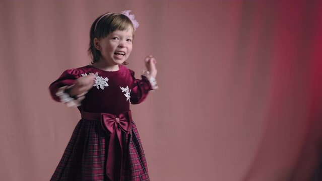 Cute little girl in an vintage burgundy dress is dancing, rejoicing and making funny faces. The concept of children theater productions and developing acting circles. Pink background