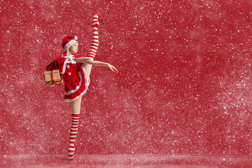 Dancing ballerina girl in pointe shoes with a gift in her hands dressed as Santa Claus on a red...