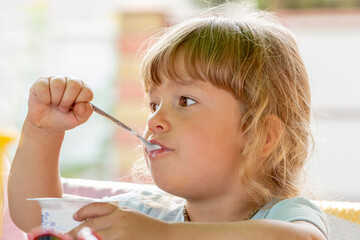 A little girl eats baby yogurt with a spoon on a neutral background.