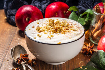 Bowl of homemade apple pie yogurt ready for breakfast, with tart crumble topping. On wooden background with red autumn apples and spices, copy space