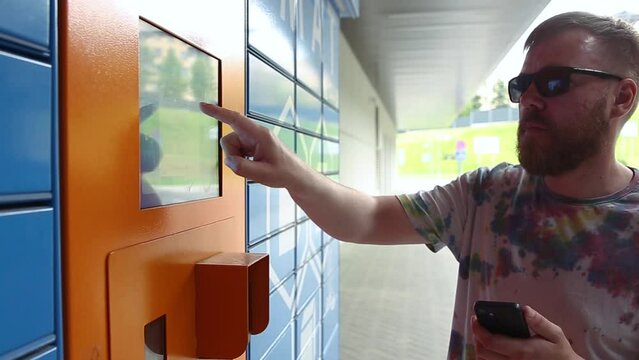 Man with smartphone enters parcel code on screen and picks up parcel from automated postal machine. Man holds smartphone using automatic self-service mail terminal closeup.