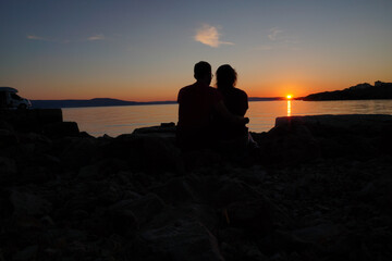 guy and girl, hugging romantic couple sitting on sea stone beach during sunset. soft light, mirroring and calm water. view from back on seashore on island, camping