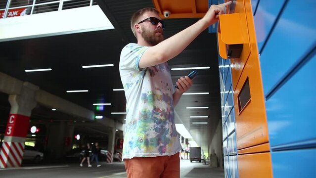 Man with smartphone enters parcel code on screen and picks up parcel from automated postal machine. Man holds smartphone using automatic self-service mail terminal closeup.
