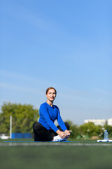 Women and sport. Girl in sportswear - blue shirt and black leggings resting on a mat on the grass in the park outdoor on a sunny day. Middle aged sportswoman dressed in sportsclothes