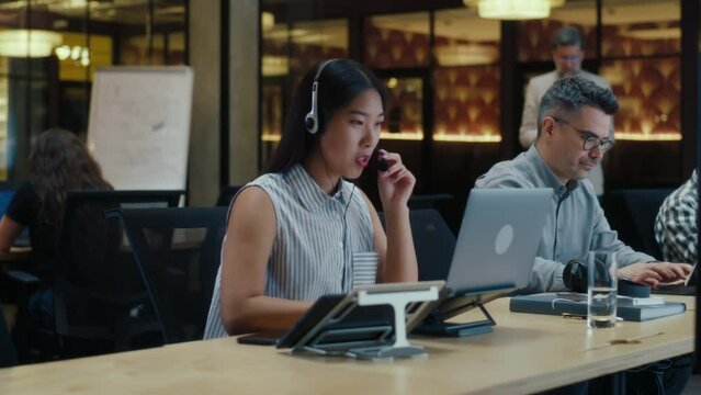 Asian female operator in headset in call center talking with client, using laptop and digital tablet. Work in call center of marketing company. Multi-ethnic office workers team