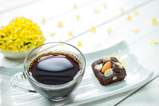 Hot americano black coffee in clear cup on white plate with chocolate brownie cake beside place on old white table and beautiful flower around