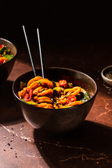 Spicy vegan Asian udon noodles stir fry with hot peppers in a moody style in a brown bowl on a...