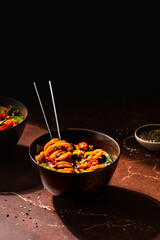 Spicy vegan Asian udon noodles stir fry with hot peppers in a moody style in a brown bowl on a brown marble table with a hint of light