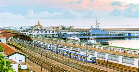 Algiers train station and port with a train parked and a coastguard Algerian Naval Forces vessel moored. The admiralty lighthouse and boats in golden hour sunlight cloudy sky.