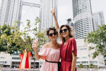 Blithesome girls in modern attire posing with skyscrapers on background. Outdoor shot of carefree ladies enjoying vacation.