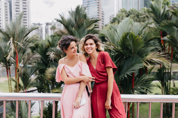 Brunette girl in pink dress spending time outdoor with best friend. Cheetful young woman posing with sister with palm trees and skyscrapers on background.