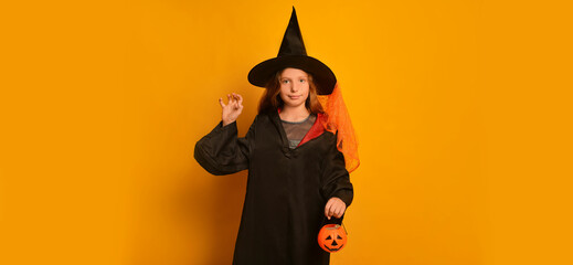 Witch girl in wizard student costume making Boo gesture, holding Halloween pumpkin candy basket or Jack o Lantern candy bucket with sweets and looking at camera on yellow background with copy space.