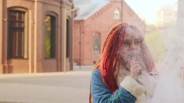 Portrait of young woman with dreadlocks vaping, standing on street. Female with colourful hairstyle smoking e-cigarette, letting off steam.