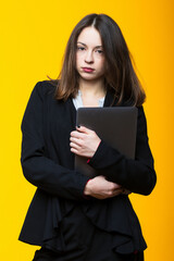 Portrait of a young woman in a suit and white shirt holding a laptop on a yellow background.