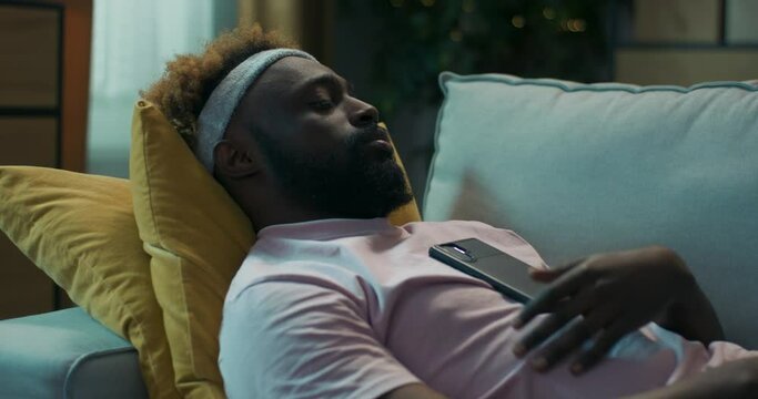 Tired young man of African appearance lay down on sofa and fell asleep on yellow pillows. Friend called him on video call. The guy woke up and started talking to friend. He has bandage on head.