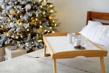 A wooden tray stands on the bed against the backdrop of Christmas lights. On the tray is a jar of cookies and two cups.