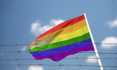 LGBTQ flag waving in the breeze inside a prison with a barbed wire fence and a blue sky with clouds.