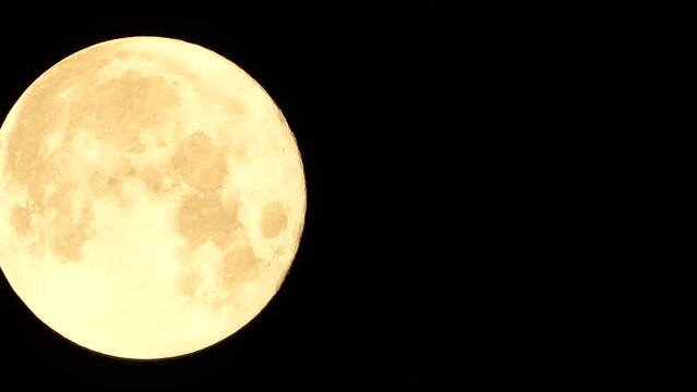 A glowing golden huge full moon seen from earth through the atmosphere against a starry night sky. A large full moon moves across the sky, the moon moving from the left frame to the right.