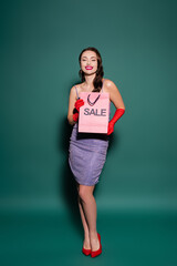 happy young woman in red gloves and purple dress holding shopping bag with sale lettering on green.