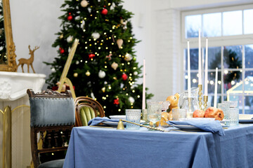 The festive dinner table stands against the backdrop of a Christmas tree. On the table there is a blue tablecloth, plates, glasses and decor