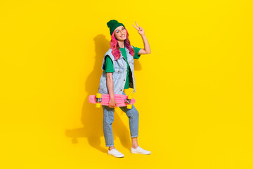 Full length photo of funky sweet lady wear jeans waistcoat spectacles showing v-sign holding skateboard isolated yellow color background