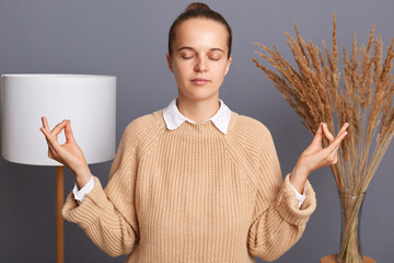Relaxed woman wearing beige jumper posing in room at home standing with raised arms and doing yoga...