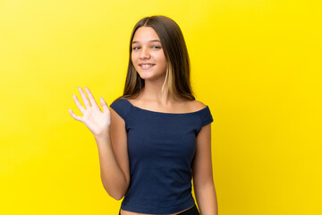 Little caucasian girl isolated on yellow background saluting with hand with happy expression