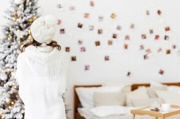 A girl in a white knitted dress and a hat stands with her back to the camera and looks at the Christmas tree