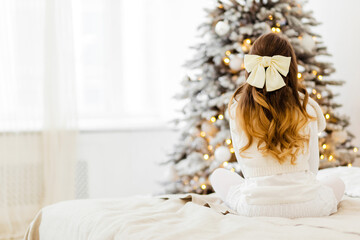A girl in a white knitted dress with hairpins in the form of a bow sits on the bed with her back to...