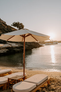 Tranquil beach scene with wooden sun loungers and white parasols lined along a pebbled shore. The turquoise sea, lush green mountains, and anchored yachts create a serene luxury escape in T&uuml;rkiye