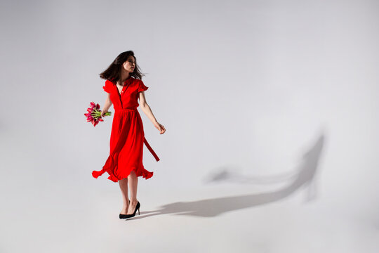 Full lengt view of a brunette young woman in long flying red dress holding a bouquet of tulips and spins, isolated grey background.