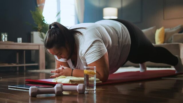 Young overweight woman doing a plank exercise having an online class with a personal trainer on a video call from home