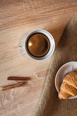 A cup of hot fragrant black coffee with a croissant in a saucer on a cloth, and cinnamon sticks, on a light brown wooden table.