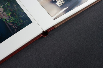 A fragment of a stylish photo book with open pages, with a leather cover in burgundy and brown colors, hardcover, lies on a gray tabletop in the room.
