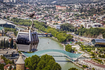 Close-up of Aerial Cable Car connecting Rike Park on the left bank of the Mtkvari river with Narikala Fortress in Tbilisi Georgia - river and Peace Bridge and busy city below - obrazy, fototapety, plakaty