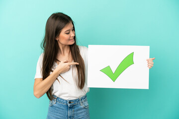 Young caucasian woman isolated on blue background holding a placard with text Green check mark icon and  pointing it