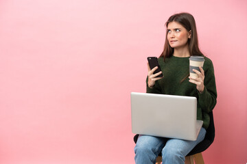 Naklejka premium Young caucasian woman sitting on a chair with her laptop isolated on pink background holding coffee to take away and a mobile while thinking something